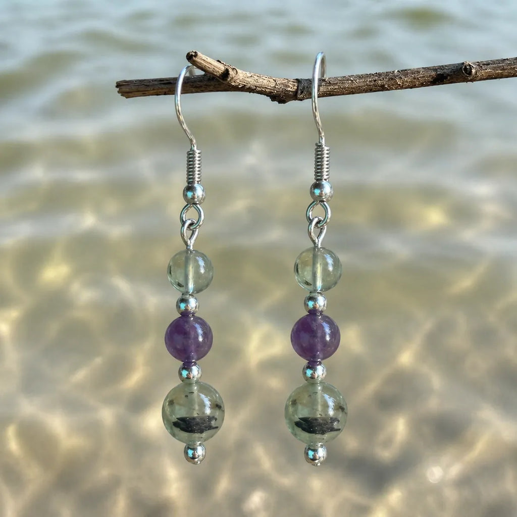 Gentle Boundaries Earrings with prehnite and amethyst gemstones on silver hook hanging on branch by the sea.