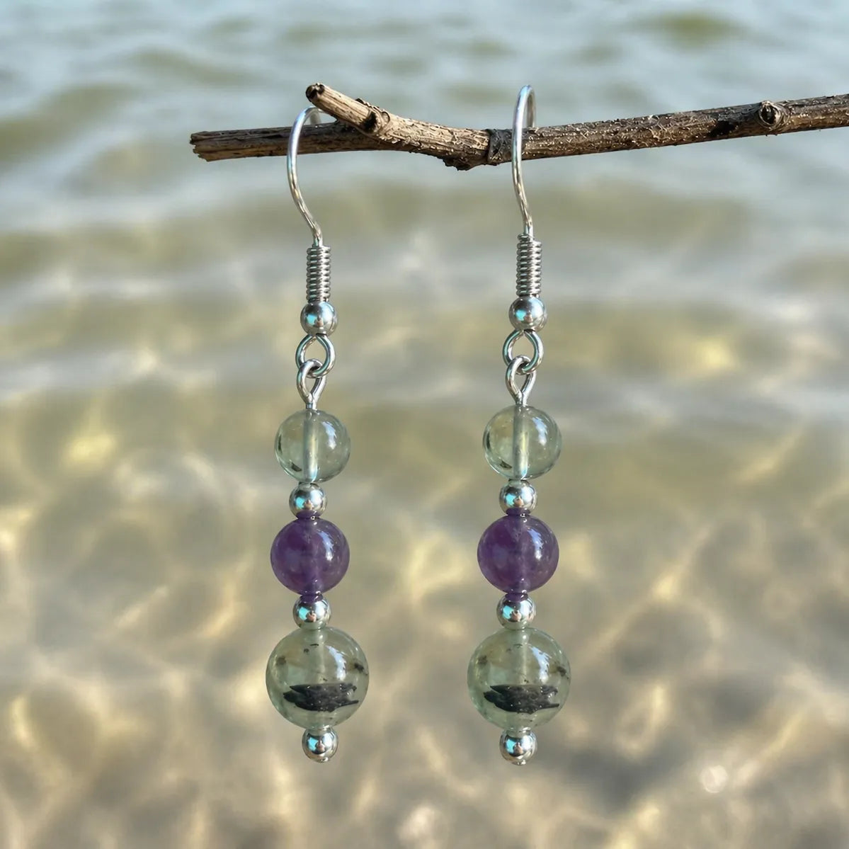 Gentle Boundaries Earrings with prehnite and amethyst gemstones on silver hook hanging on branch by the sea.