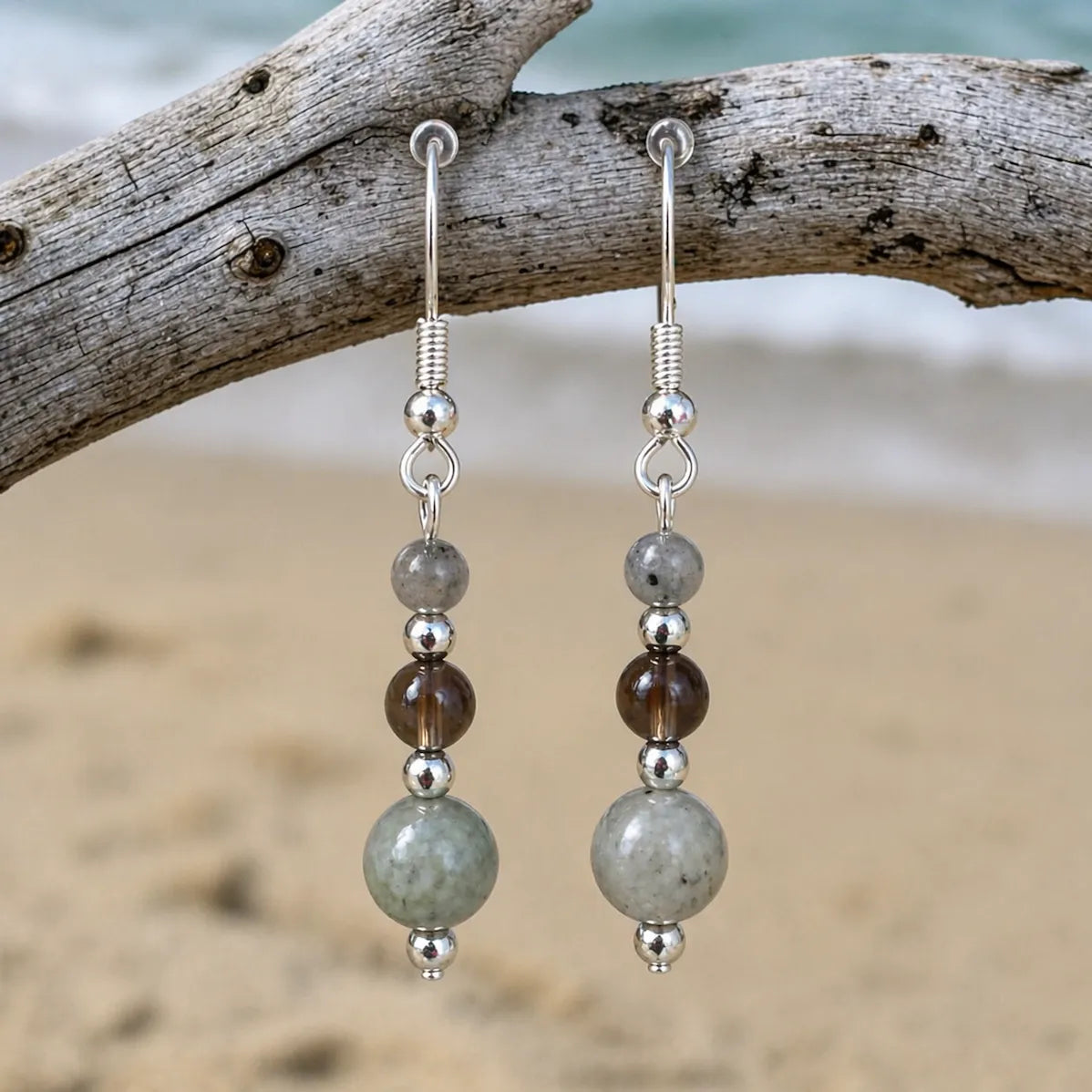 Grounded Strength Earrings with labroradite and smokey quartz gemstones on silver hook hanging straight on driftwood by the beach.