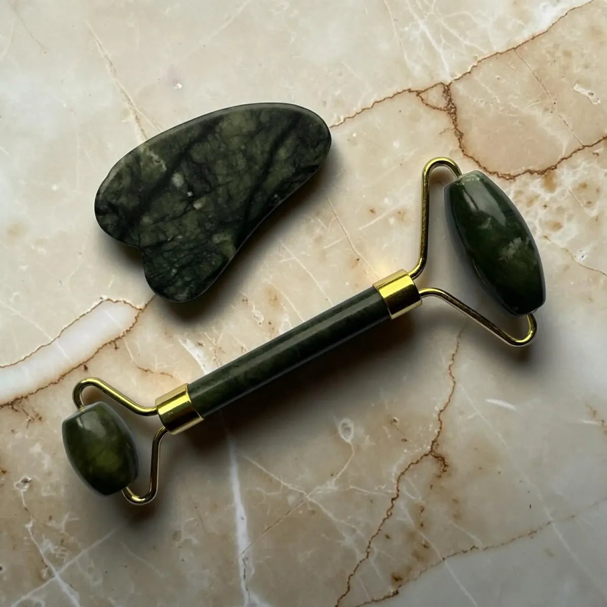 Green jade gua sha roller and stone on a marble surface.