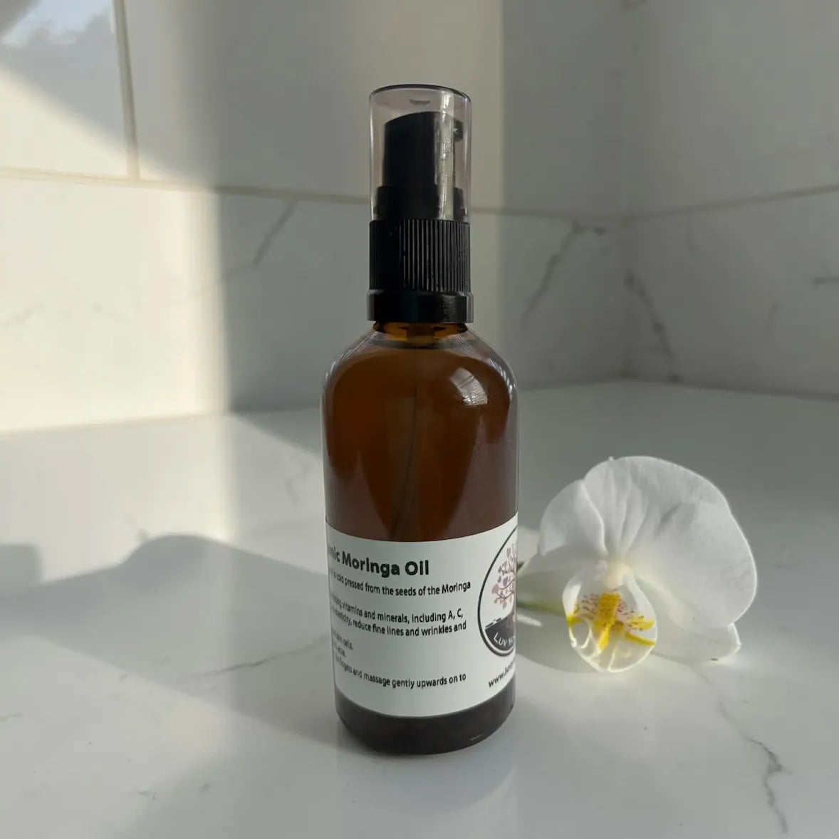 Brown bottle of moringa oil with black spray nozzle on a white surface with a white flower.