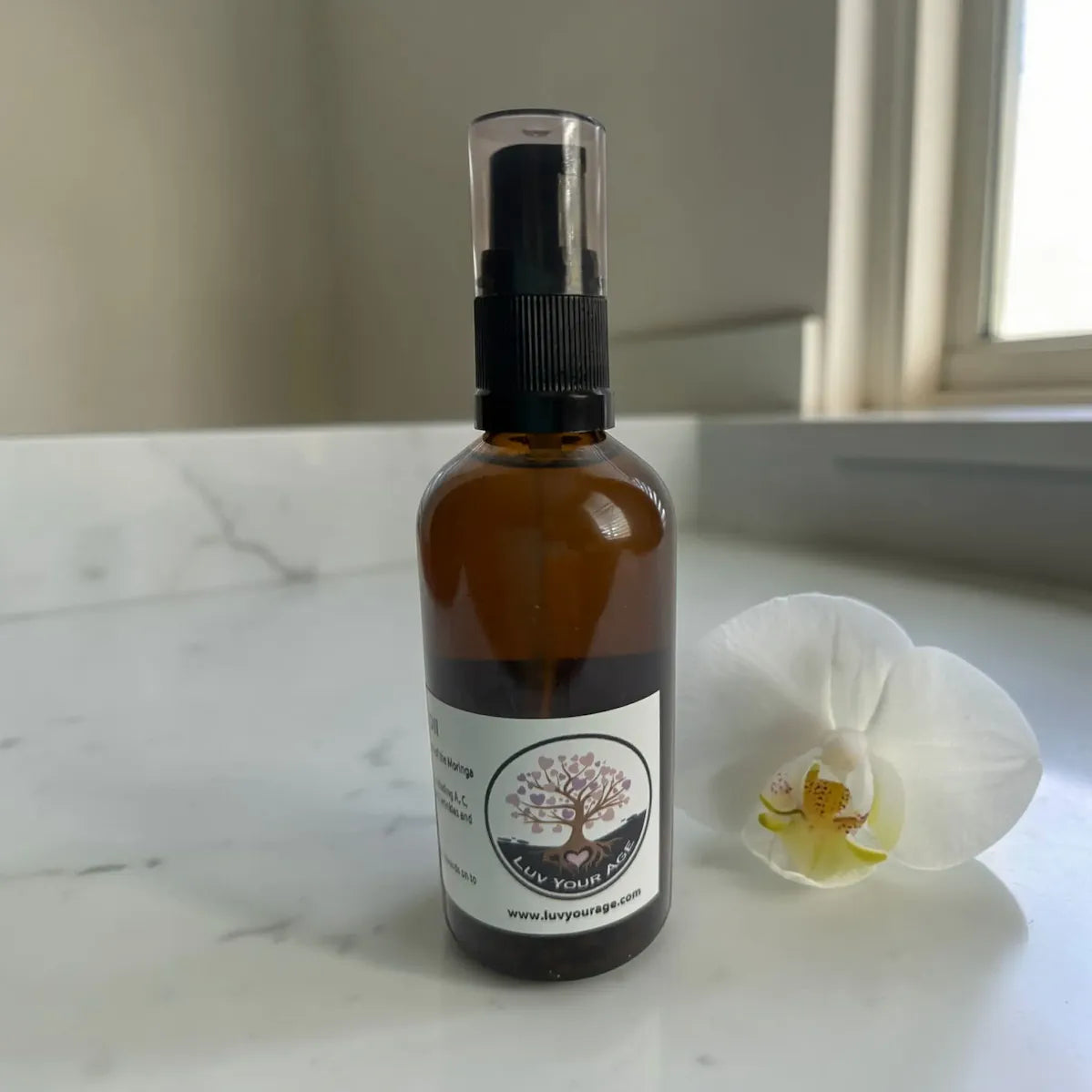 Brown spray bottle of moringa oil with a label next to a white flower on a marble surface.